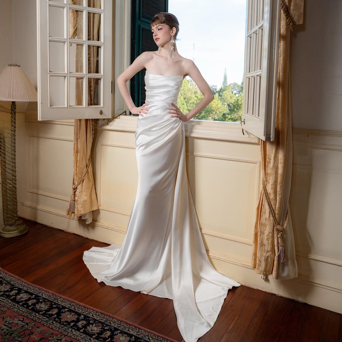 May include: A strapless, ivory-colored wedding gown with a draped bodice and a flowing, floor-length skirt. The dress features a subtle train and is made of a smooth, satin-like fabric. The model is standing near a window.