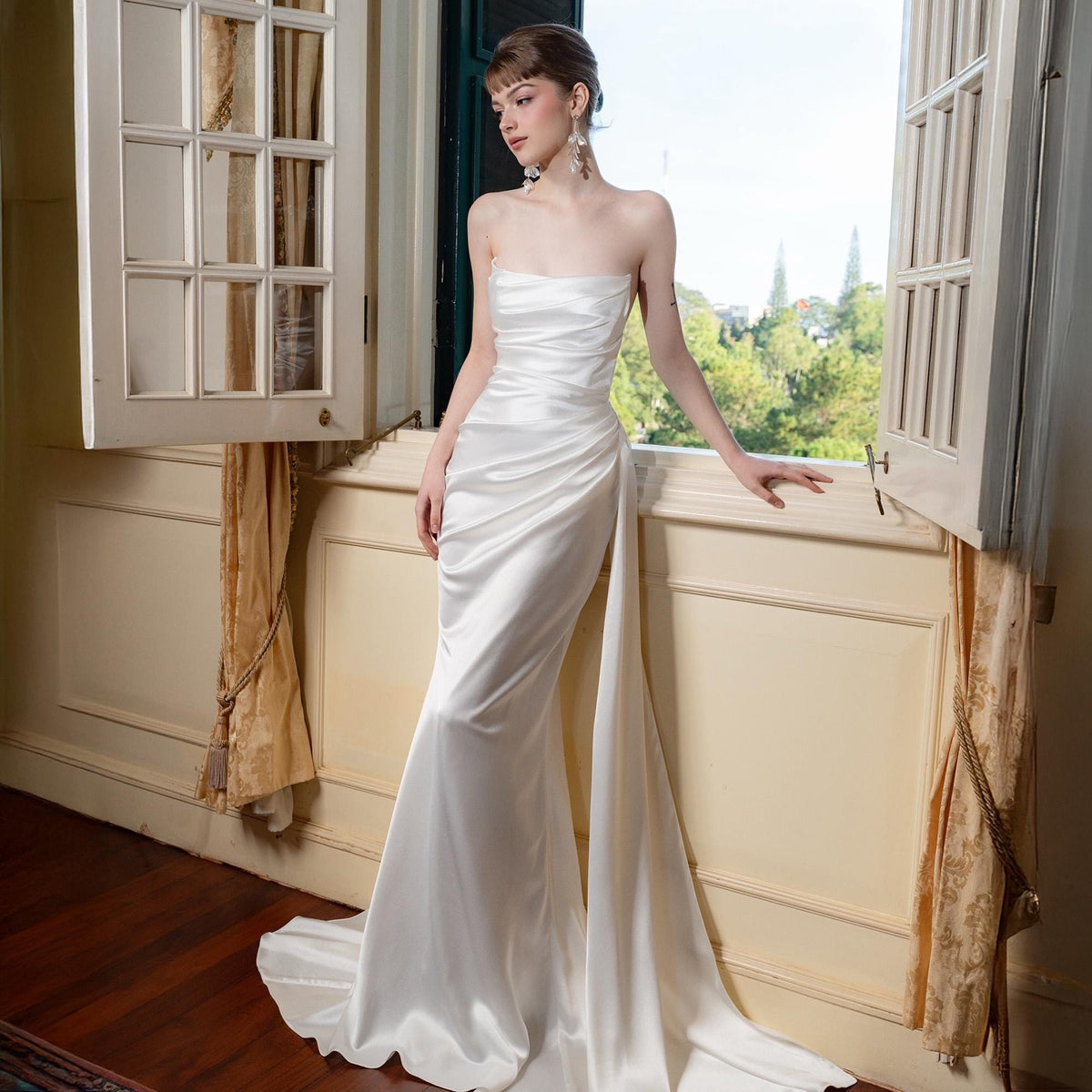 May include: A strapless, white satin wedding gown with a mermaid silhouette. The dress features a draped bodice and a flowing skirt with a side panel that extends into a train. The gown is elegant and suitable for a formal event.