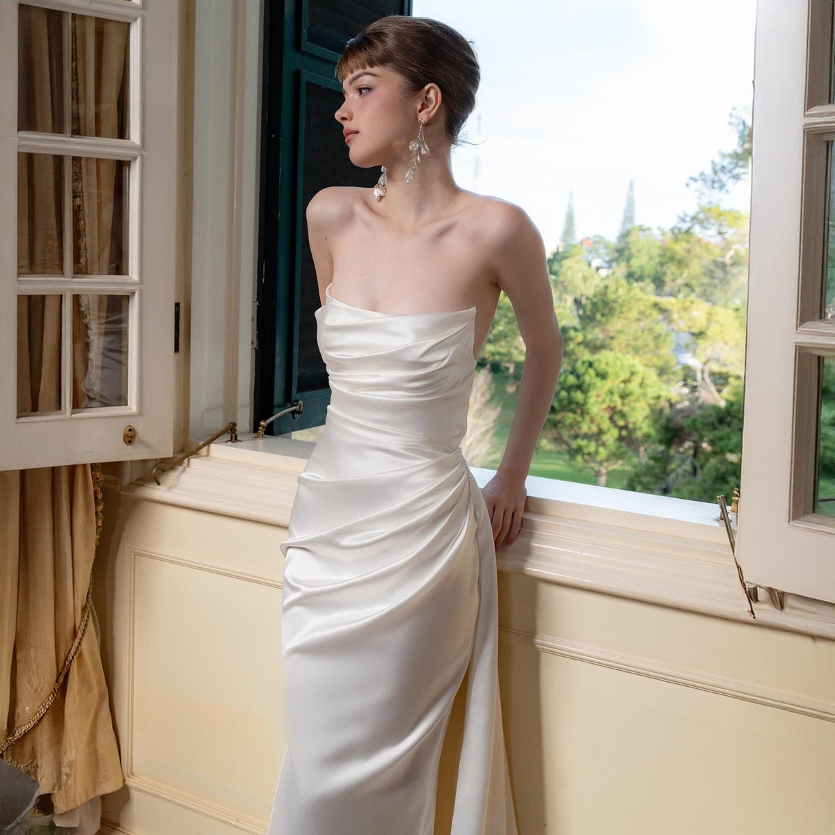 May include: A strapless, ivory-colored satin gown with a draped bodice and a flowing skirt. The dress features a side slit and is worn by a person standing near a window with a view of trees. The person is wearing dangling earrings.