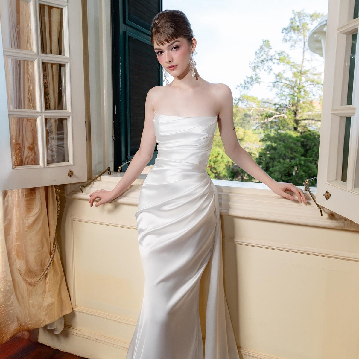 May include: A strapless white satin gown with a fitted bodice and a flowing skirt. The dress features elegant draping and a subtle train. The model is standing near an open window with a view of trees.