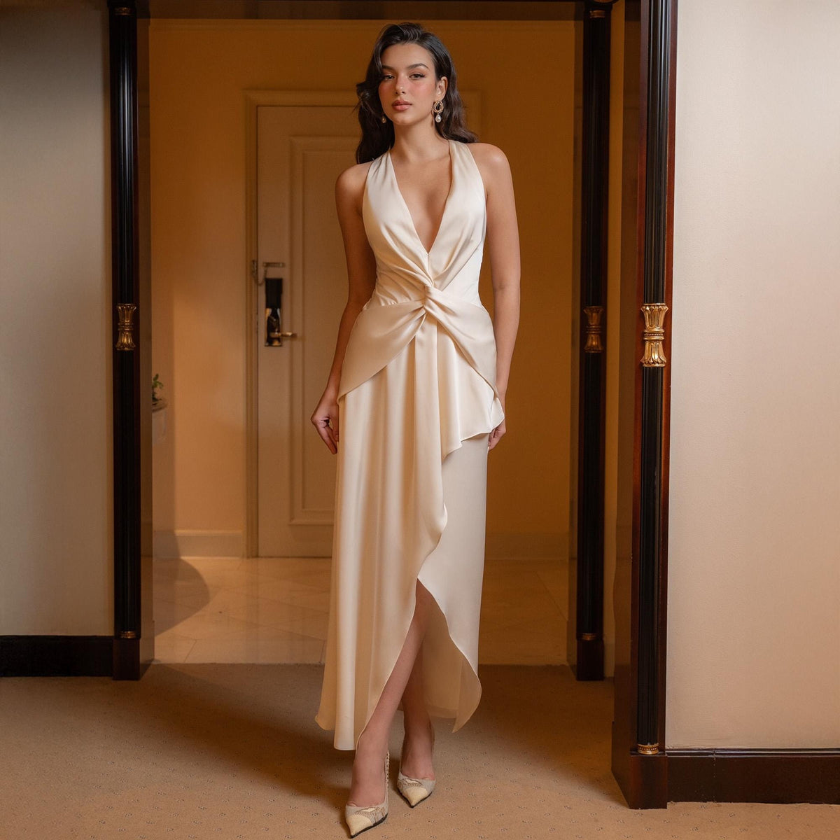 May include: A cream-colored halter neck dress with a plunging neckline and a flowing, asymmetrical skirt. The dress features a twisted detail at the waist and is paired with matching heels. The model is standing in a doorway.