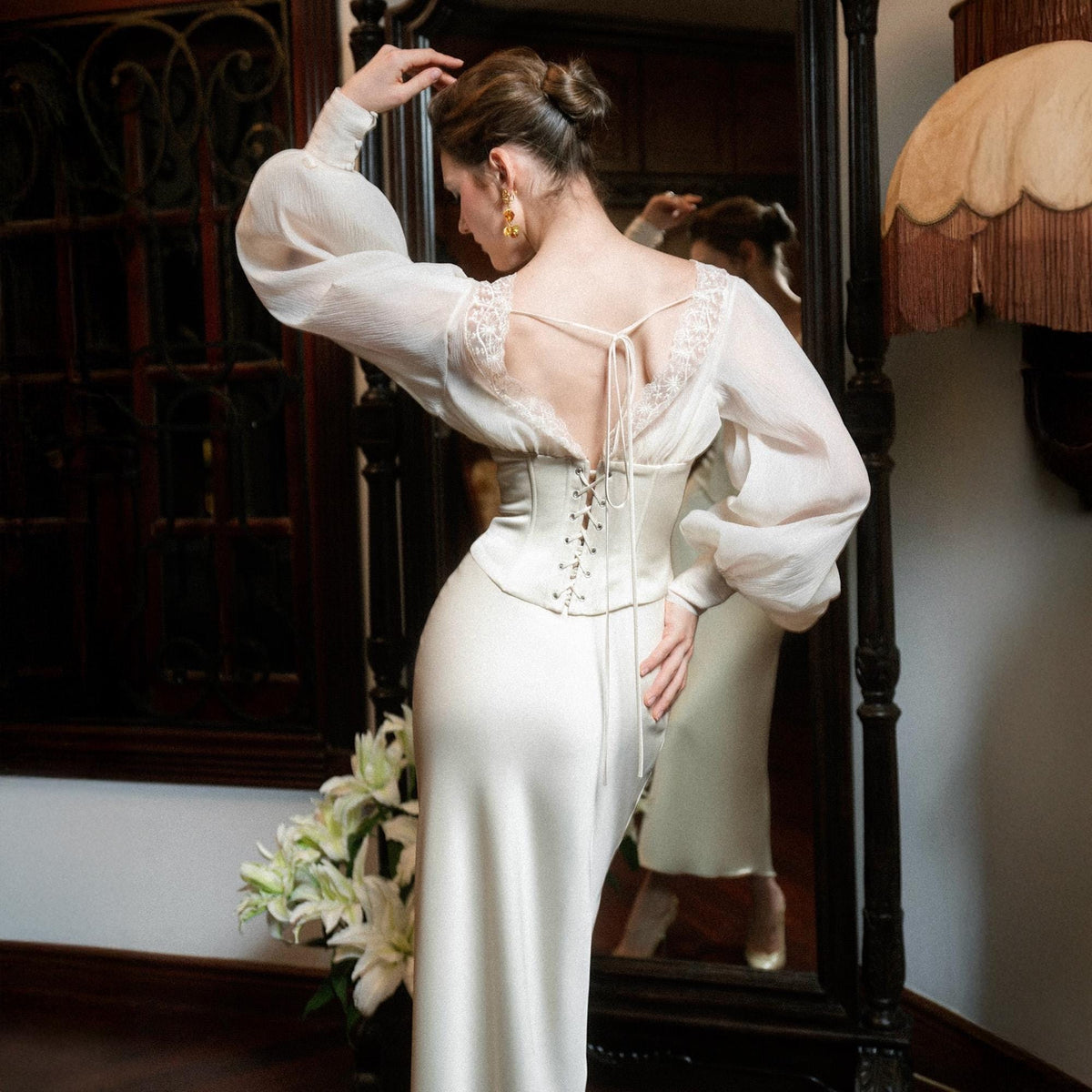 May include: A white satin dress with a corset bodice and a long skirt. The dress features a sheer, long-sleeved top with lace detailing and a low back. The model is wearing gold earrings. The dress is elegant and suitable for special occasions.