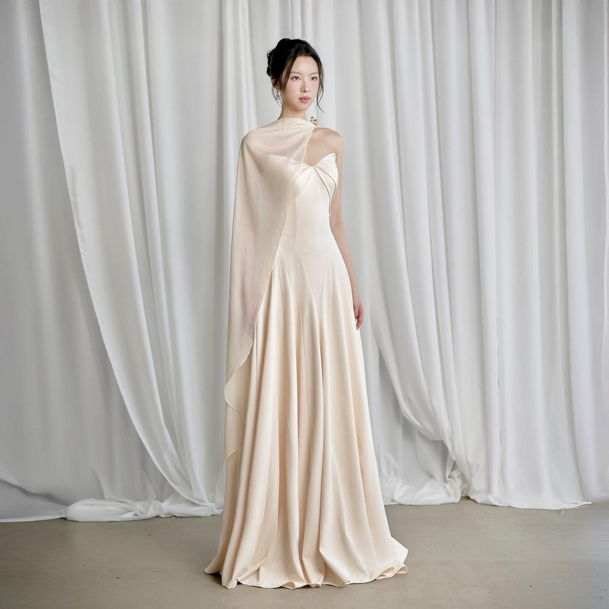 May include: A floor-length, cream-colored gown with a draped cape. The dress features a fitted bodice and a flowing skirt. The cape is attached at the shoulder and drapes over the arm. The background is a white curtain.
