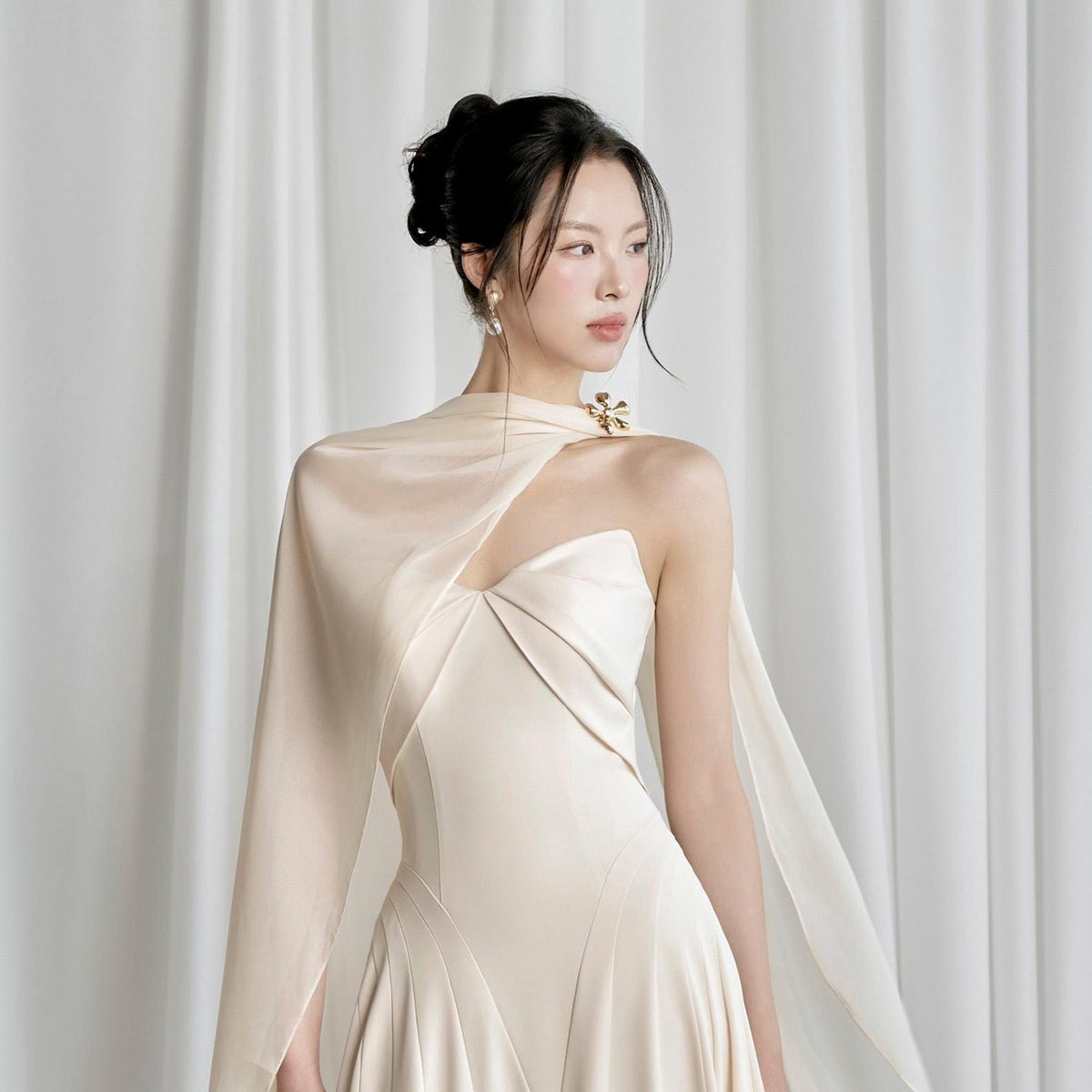 May include: A cream-colored formal gown with a draped cape detail. The dress features a strapless bodice and a flowing skirt. A gold-toned brooch accents the shoulder. The model has dark hair styled in a bun.