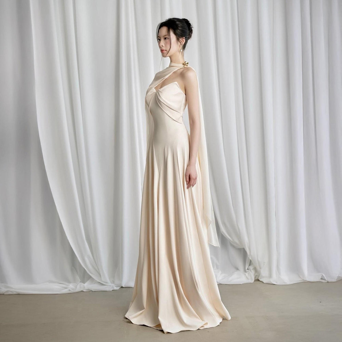 May include: A floor-length, cream-colored gown with a draped bodice and a flowing skirt. The dress features a one-shoulder design with a cape-like overlay. A gold-colored embellishment adorns the shoulder. The fabric has a satin-like sheen.