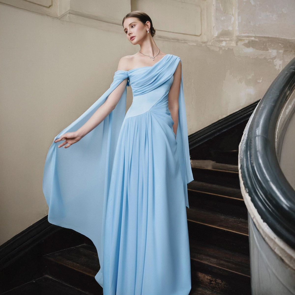 May include: A light blue, one-shoulder gown with flowing cape sleeves. The dress features a fitted bodice and a full, floor-length skirt. The model is standing on a wooden staircase with a dark banister. The dress is made of a soft, draping fabric.