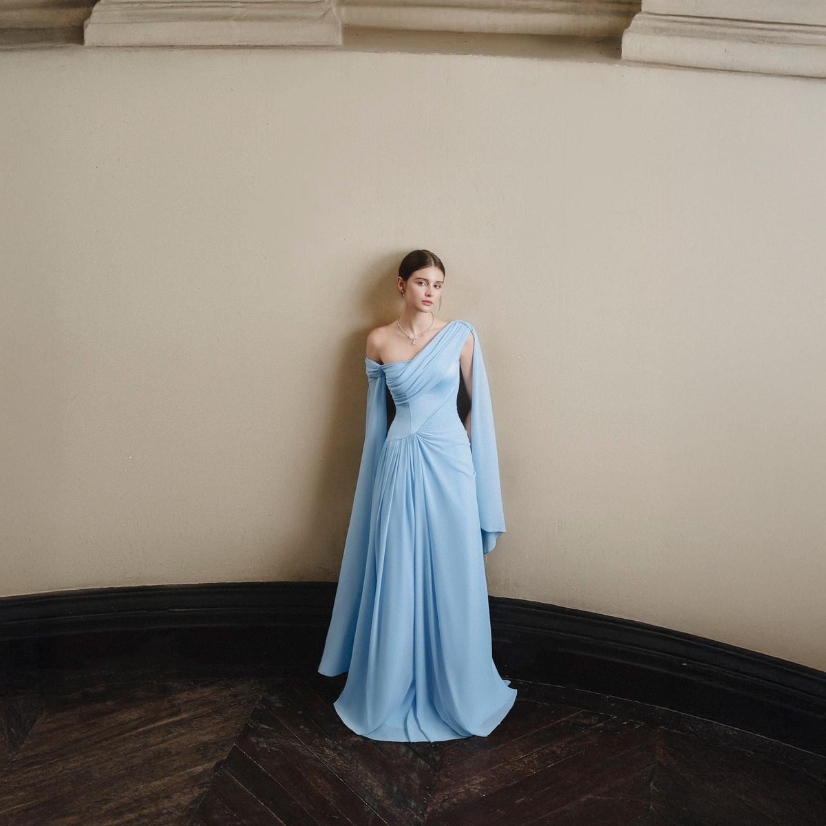 May include: A light blue, floor-length gown with an asymmetrical neckline and flowing sleeves. The dress features a draped bodice and a pleated skirt. The model is standing in a room with a light-colored wall and dark wood floor.