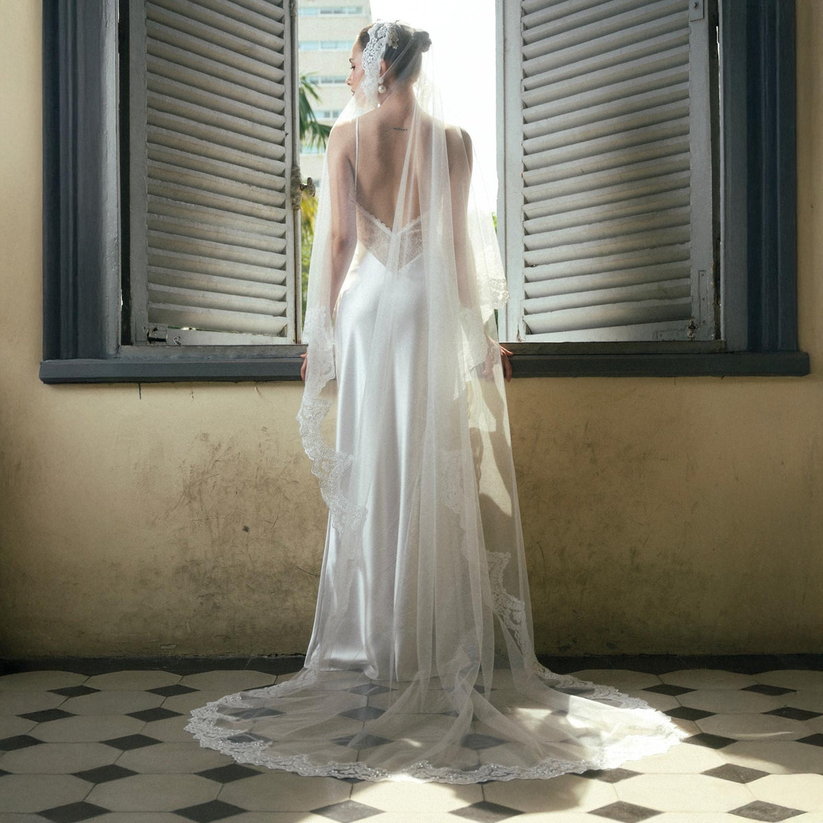 May include: A white wedding dress with a low back and a long, flowing train. The dress is paired with a delicate lace veil. The bride is standing near a window with wooden shutters.