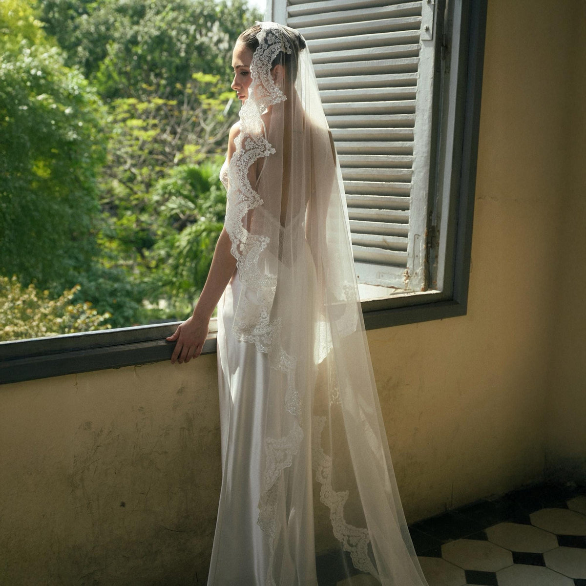 May include: A bride in a white wedding dress and veil stands near a window. The veil is long and sheer, with delicate lace trim along the edges. The dress is simple and elegant, with a flowing silhouette. The background features lush green foliage.