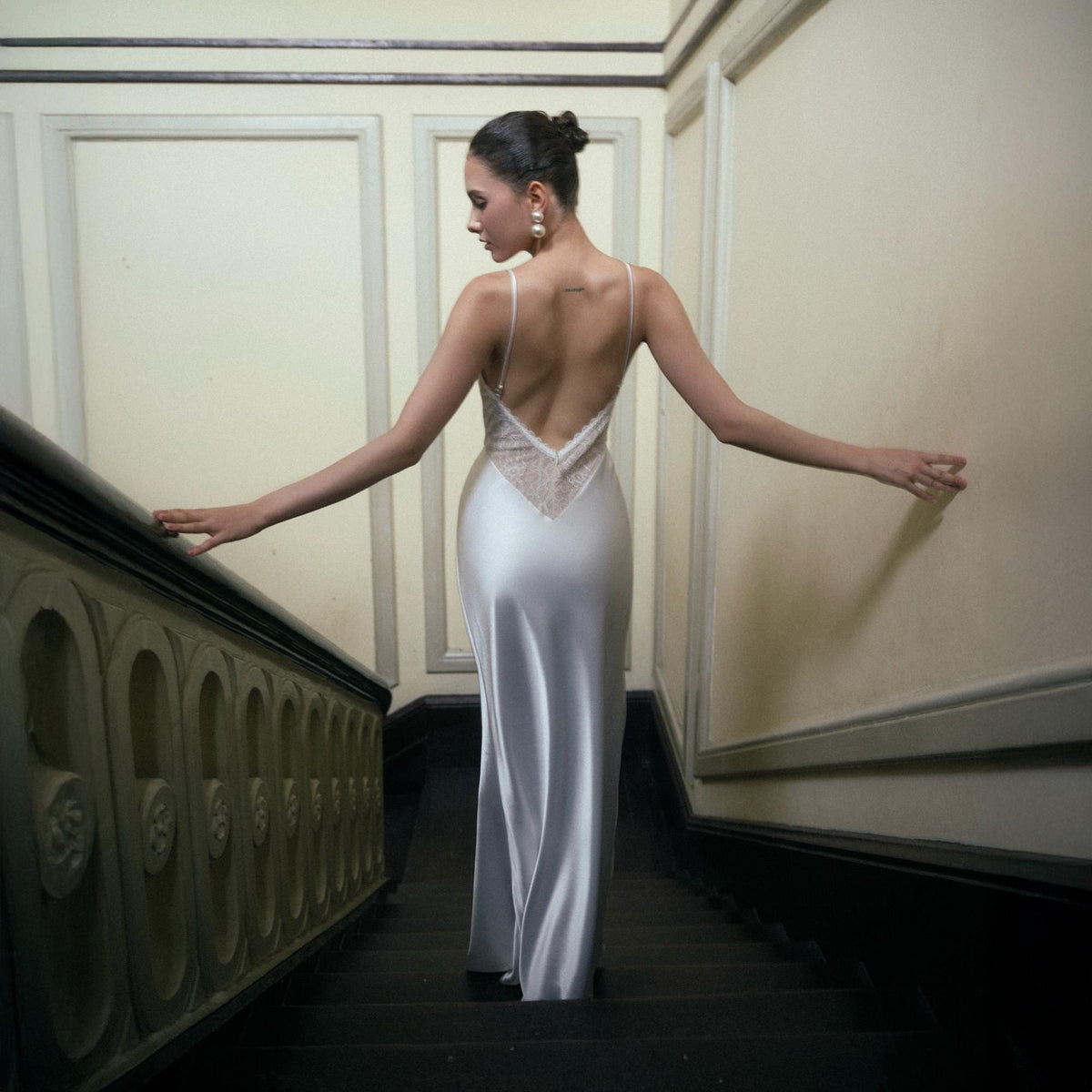 May include: A long, flowing, silver satin gown with a low, open back and delicate spaghetti straps. The back features a lace panel. The woman is standing on a staircase with a dark banister and steps.