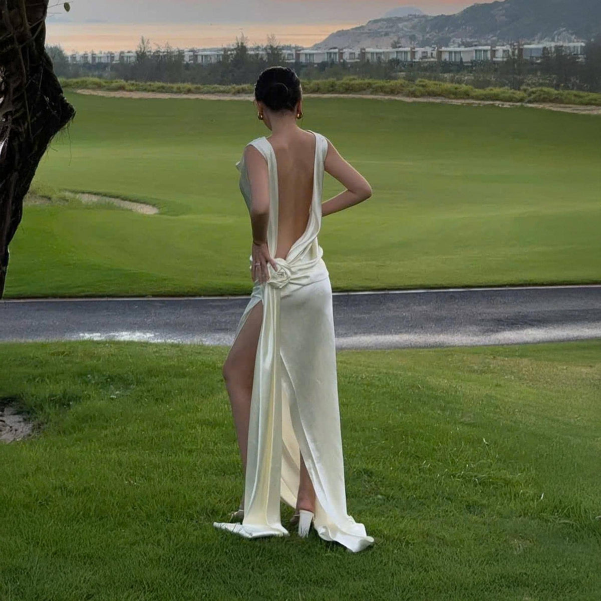 May include: A cream-colored, floor-length gown with a low back and high slit. The dress features a draped design and a floral accent at the waist. The model is standing on a green lawn.
