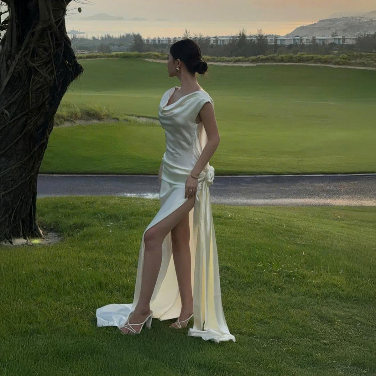 May include: A cream-colored, satin gown with a cowl neckline and a high slit. The dress features a flowing train and is accessorized with white heels. The image is set outdoors on a green lawn.