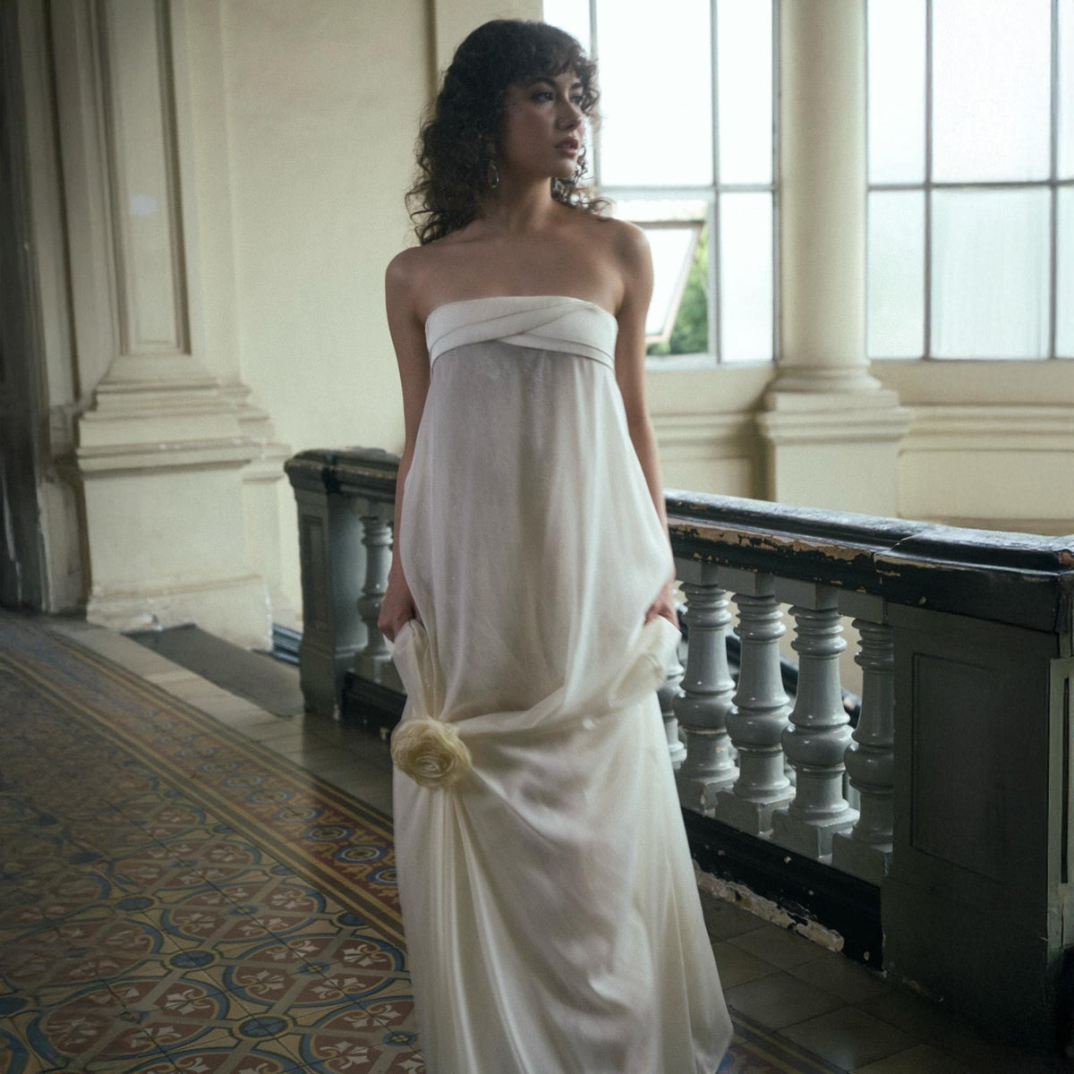 May include: A strapless ivory gown with a draped bodice and a flowing skirt. A large, cream-colored floral accent adorns the side. The dress is worn in a well-lit interior with a patterned floor and architectural details.