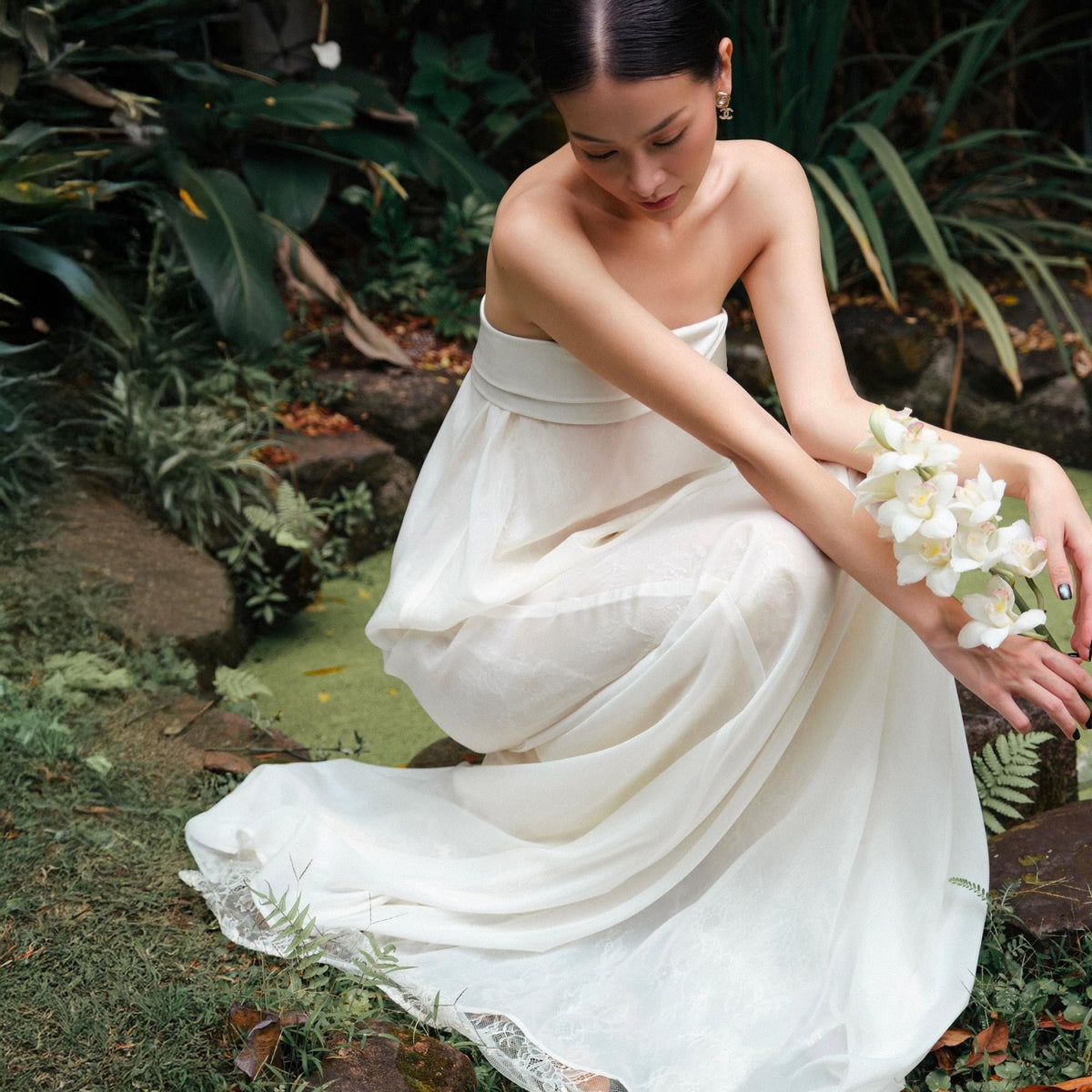 May include: A strapless, ivory-colored dress with a flowing skirt and a delicate lace trim. The dress is worn by a person in a natural outdoor setting, holding white flowers. The dress appears to be made of a lightweight, sheer fabric.