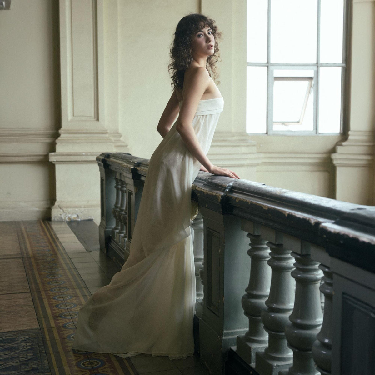 May include: A woman in a white, strapless gown stands near a dark wooden railing in a building with large windows. The dress flows to the floor, and the woman has curly brown hair. The background features columns and a patterned floor.