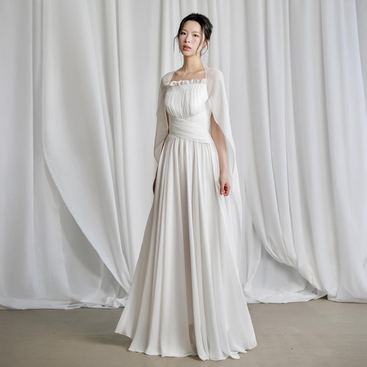 May include: A white wedding dress with a square neckline and a flowing skirt. The dress features a fitted bodice with pleating and a waist sash. A sheer, cape-like overlay drapes over the shoulders. The model is standing in front of white curtains.