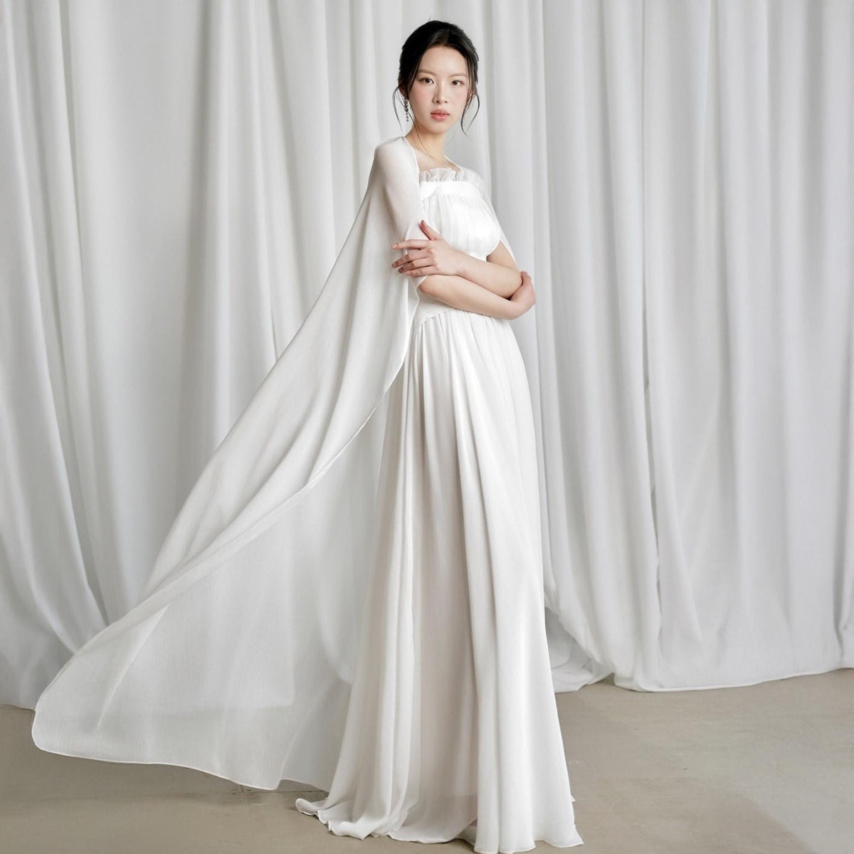 May include: A white floor-length gown with a flowing cape. The dress features a fitted bodice and a long, flared skirt. The cape drapes elegantly from the shoulders, creating a graceful silhouette. The fabric appears to be lightweight and flowing, perfect for a special occasion.