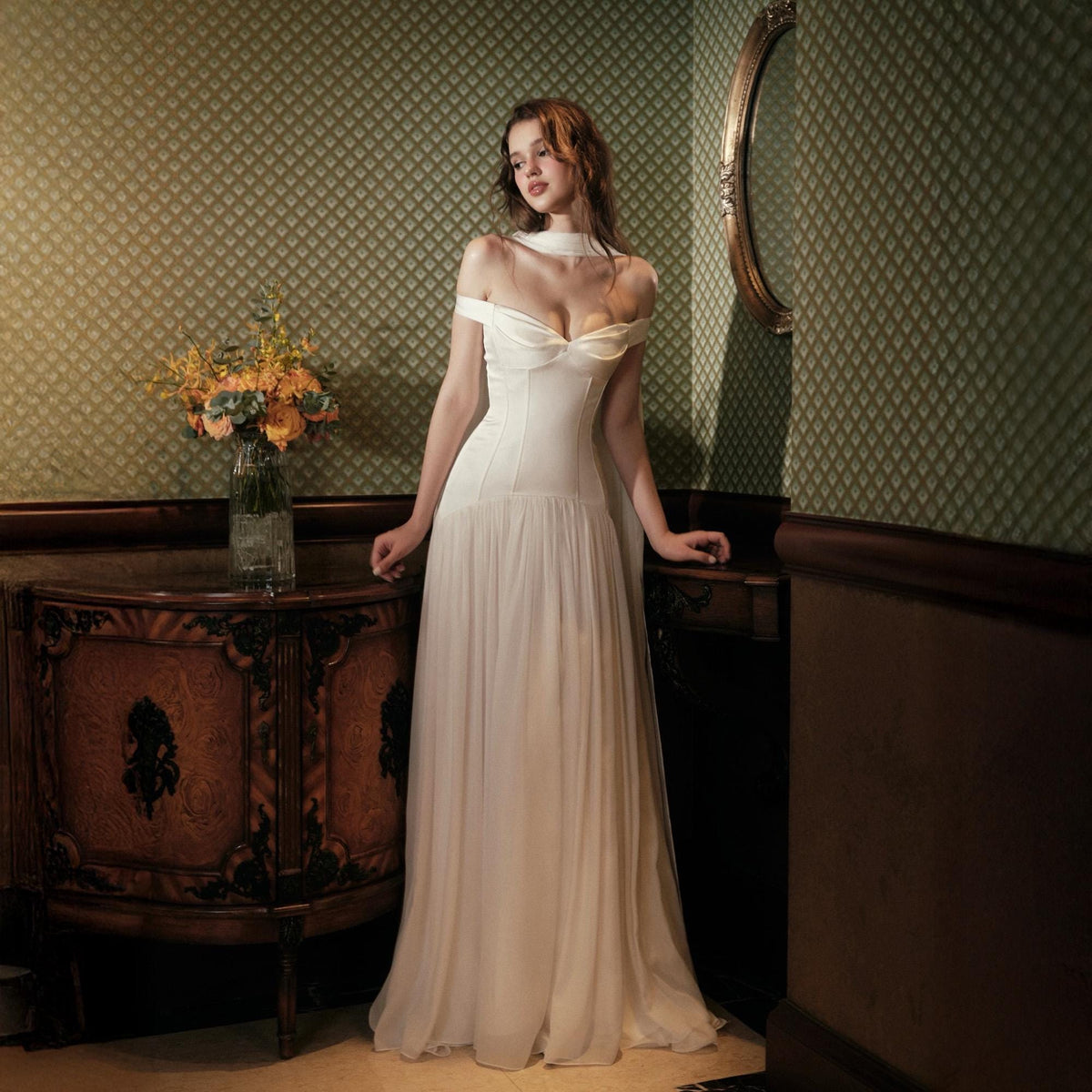 May include: A floor-length off-the-shoulder white gown featuring a structured bodice with a sweetheart neckline and a flowing, sheer skirt. The dress has a fitted waistline and a smooth, opaque fabric panel across the chest, transitioning into a soft, layered skirt that reaches the floor.