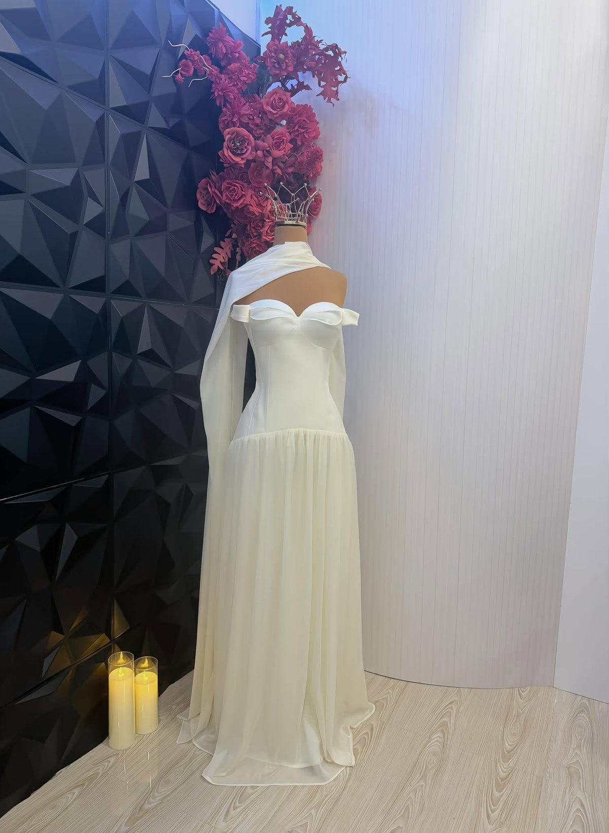 May include: A floor-length off-white gown featuring a sweetheart neckline, off-the-shoulder sleeves, and a draped fabric panel extending from the shoulder. The dress has a fitted bodice and a flowing, sheer skirt, displayed on a mannequin.