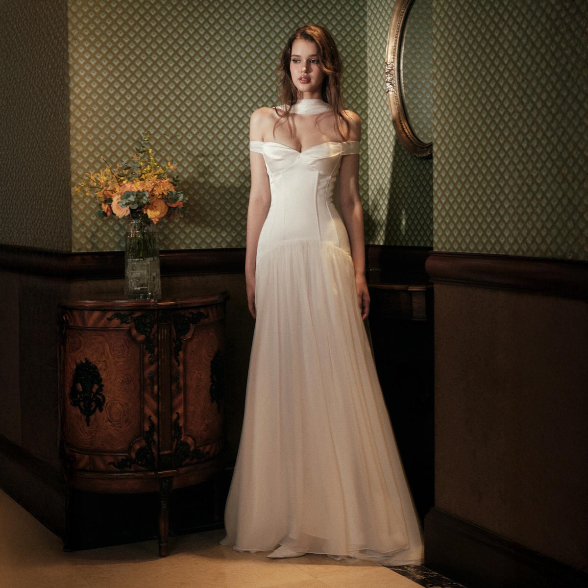 May include: A floor-length white gown featuring an off-the-shoulder neckline with a draped bodice, a fitted waistline, and a flowing, sheer skirt. The garment includes a matching neck scarf detail that rests around the collarbone area.