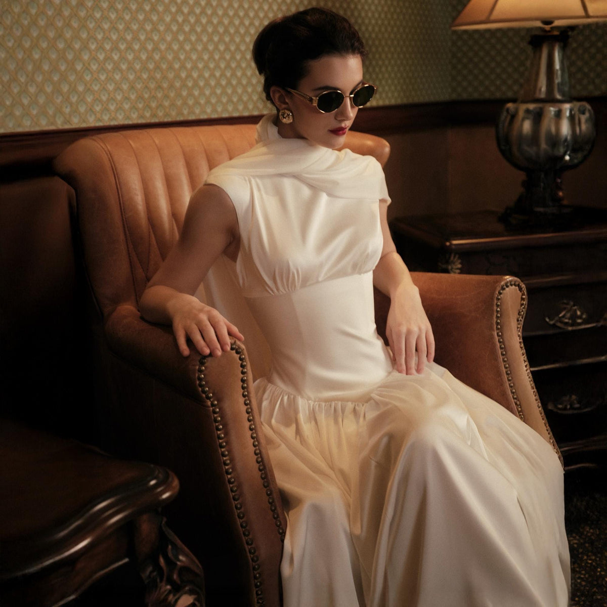 May include: A woman wearing a white satin dress with a draped neckline and a flowing skirt, seated in a brown leather armchair. She is wearing sunglasses and gold earrings. The room has a vintage aesthetic with a patterned wallpaper.