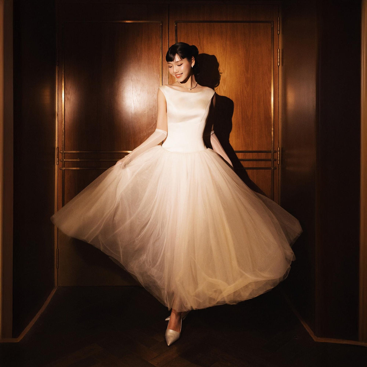 May include: A woman in a white, sleeveless dress with a full tulle skirt and satin bodice. She wears long white gloves and silver heels. The dress is illuminated by warm light, creating a soft, elegant look. The background is a dark wooden door.