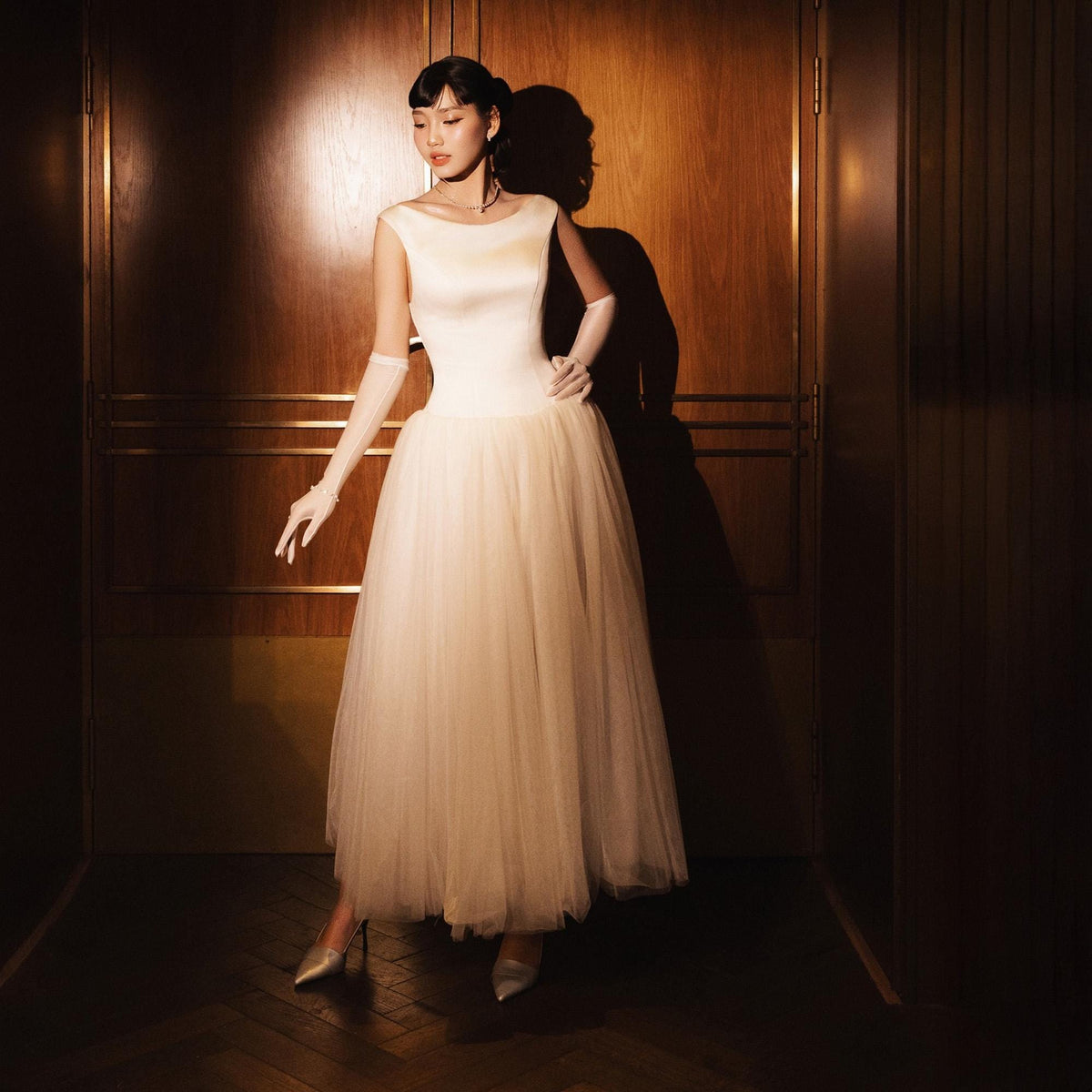 May include: A formal ivory-colored gown with a fitted bodice and a flowing tulle skirt. The dress features a bateau neckline and is paired with long white gloves and silver heels. The background is a wooden door.