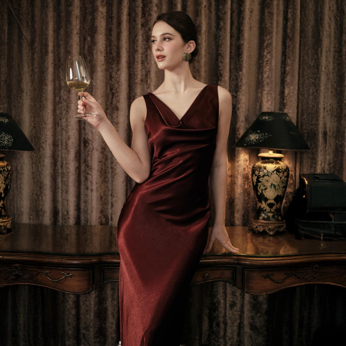 May include: A burgundy sleeveless dress with a cowl neckline. The dress is made of a silky material and has a slight sheen. The model is holding a wine glass. The background includes a wooden table and lamps.