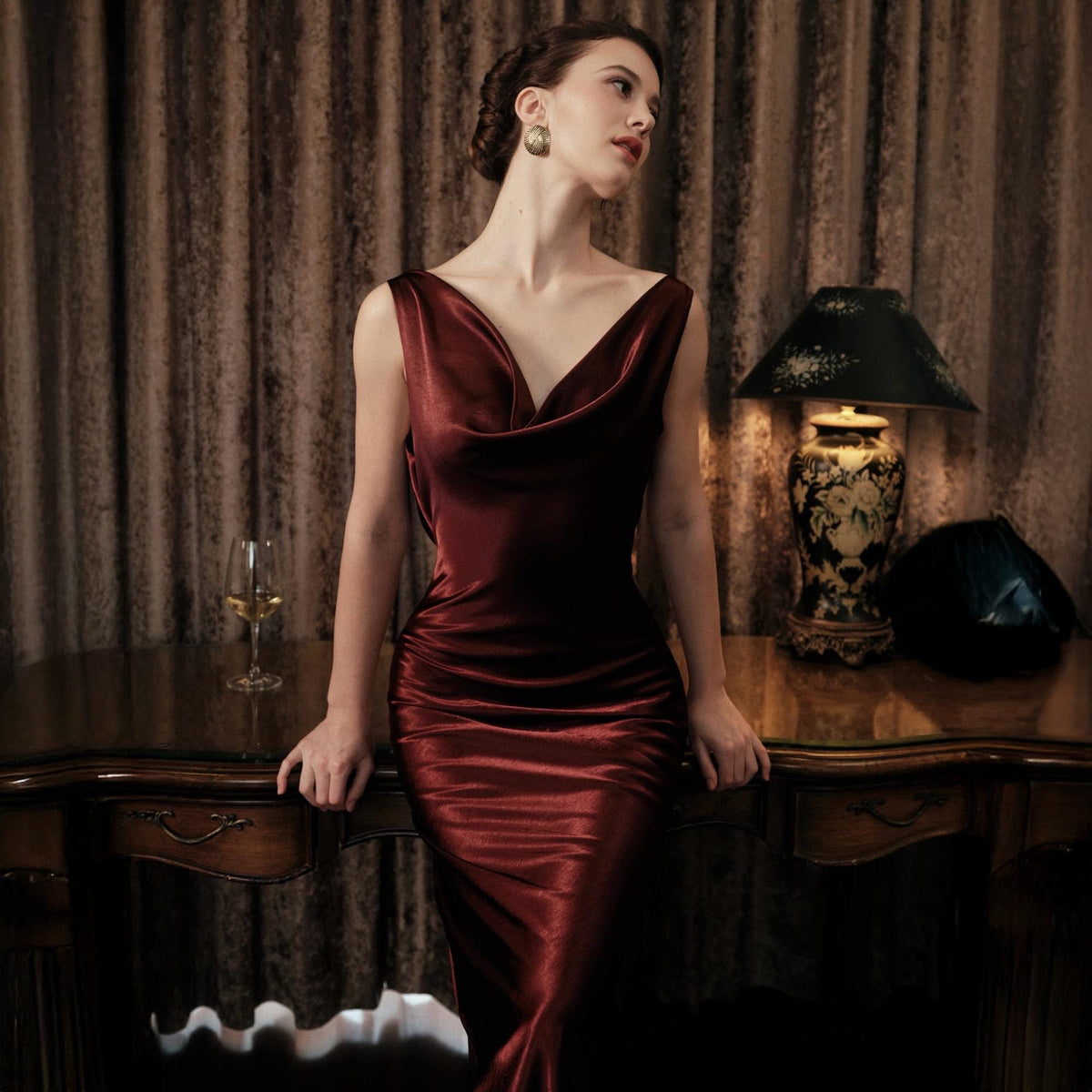 May include: A burgundy, floor-length dress with a cowl neckline. The dress is made of a shiny, silky material and has a fitted silhouette. The woman is wearing gold earrings and is leaning on a wooden table.
