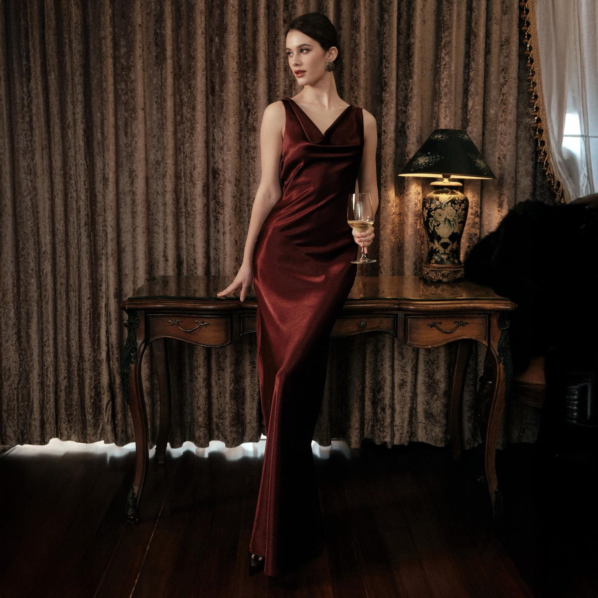 May include: A floor-length, burgundy velvet gown with a cowl neckline. The dress is sleeveless and form-fitting, with a slight train. The woman is holding a glass of white wine. The dress is elegant and suitable for formal events.