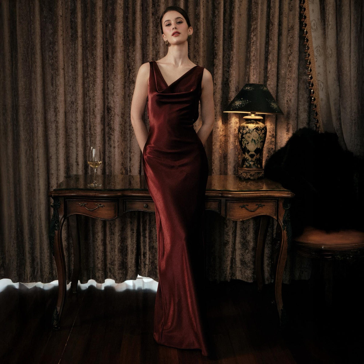 May include: A burgundy-colored, sleeveless, floor-length dress with a cowl neckline. The dress is made of a silky material and is worn by a person. A vintage-style desk, lamp, and chair are in the background.