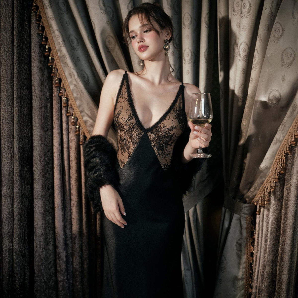 May include: A black satin slip dress with a lace bodice and spaghetti straps. The dress features a plunging neckline and a flowing skirt. A glass of white wine is held in the right hand. A black faux fur sleeve is on the left arm.