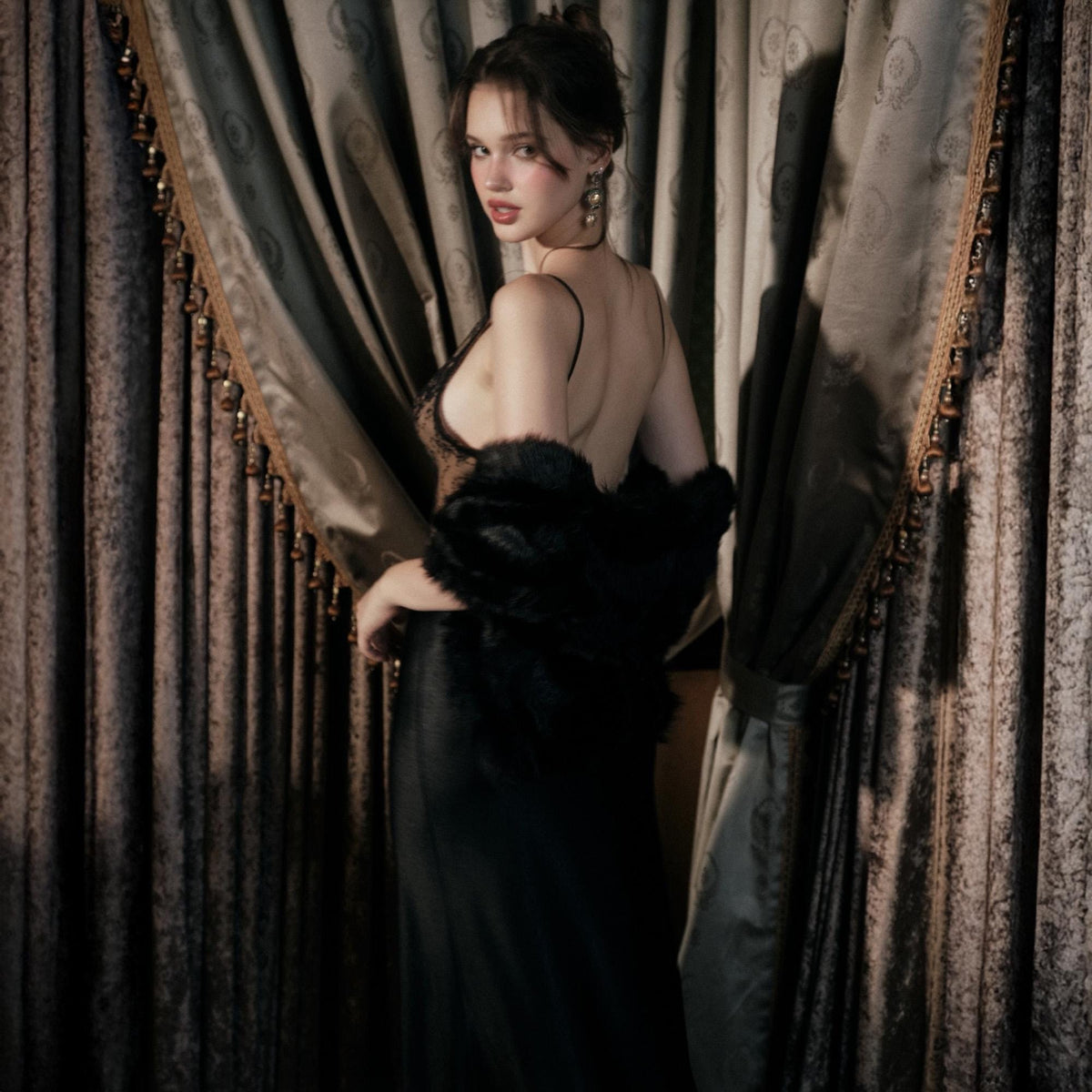May include: A woman in a black evening gown with spaghetti straps and a fur wrap. The dress has a low back and a long skirt. The woman is looking over her shoulder. The background features ornate curtains.