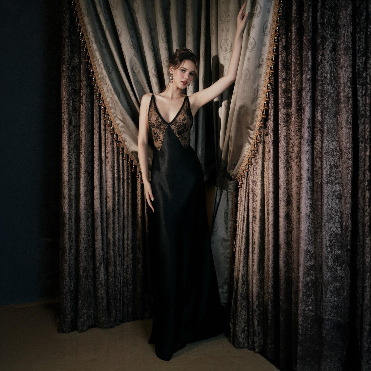 May include: A black satin slip dress with a V-neckline and lace detailing on the bodice. The dress is floor-length and has thin straps. The model is standing in front of a dark curtain.