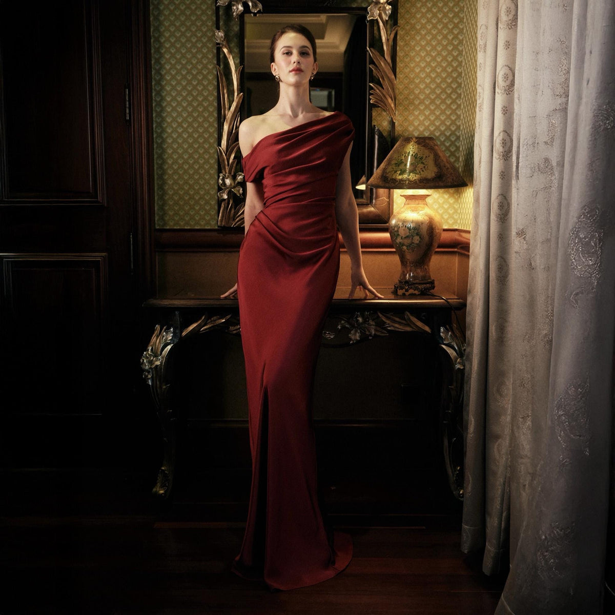 May include: A floor-length, one-shoulder red gown with a draped bodice and a high slit. The dress is worn in a room with a decorative mirror, a table, and a lamp.
