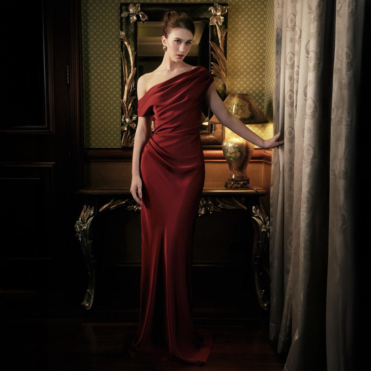 May include: A formal, floor-length, one-shoulder red dress. The dress features draped fabric across the bodice and a fitted silhouette. The image is set in a room with a decorative mirror and a table with a lamp.