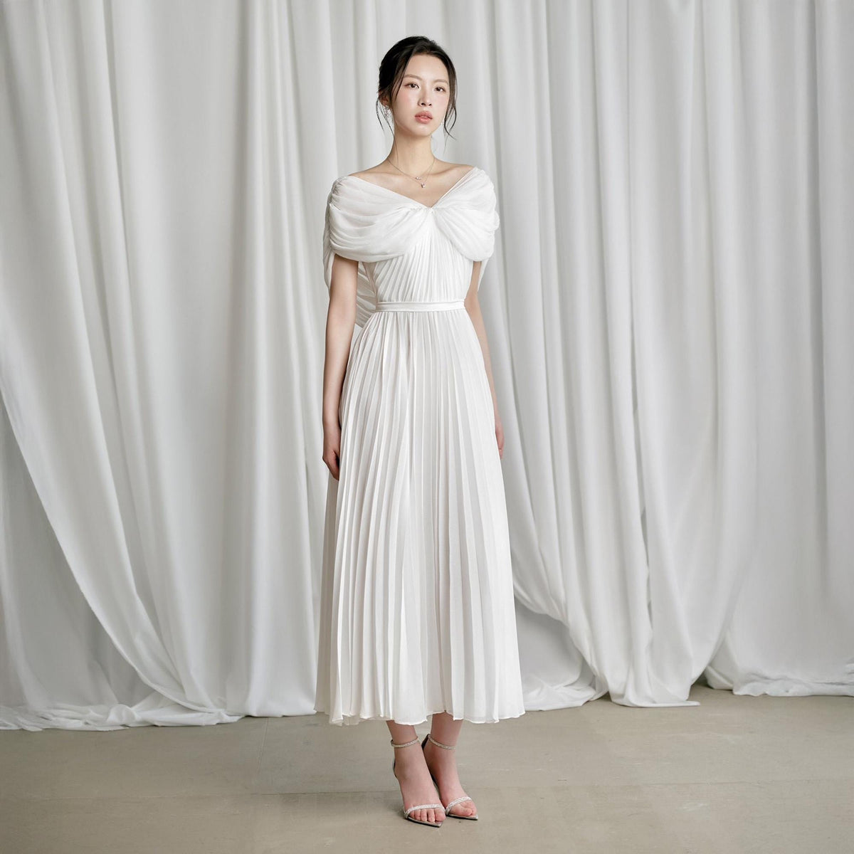 May include: A white off-the-shoulder dress with a pleated skirt. The bodice features a draped design, and the dress has a fitted waist. The model is wearing light-colored heels. The background is a white curtain.