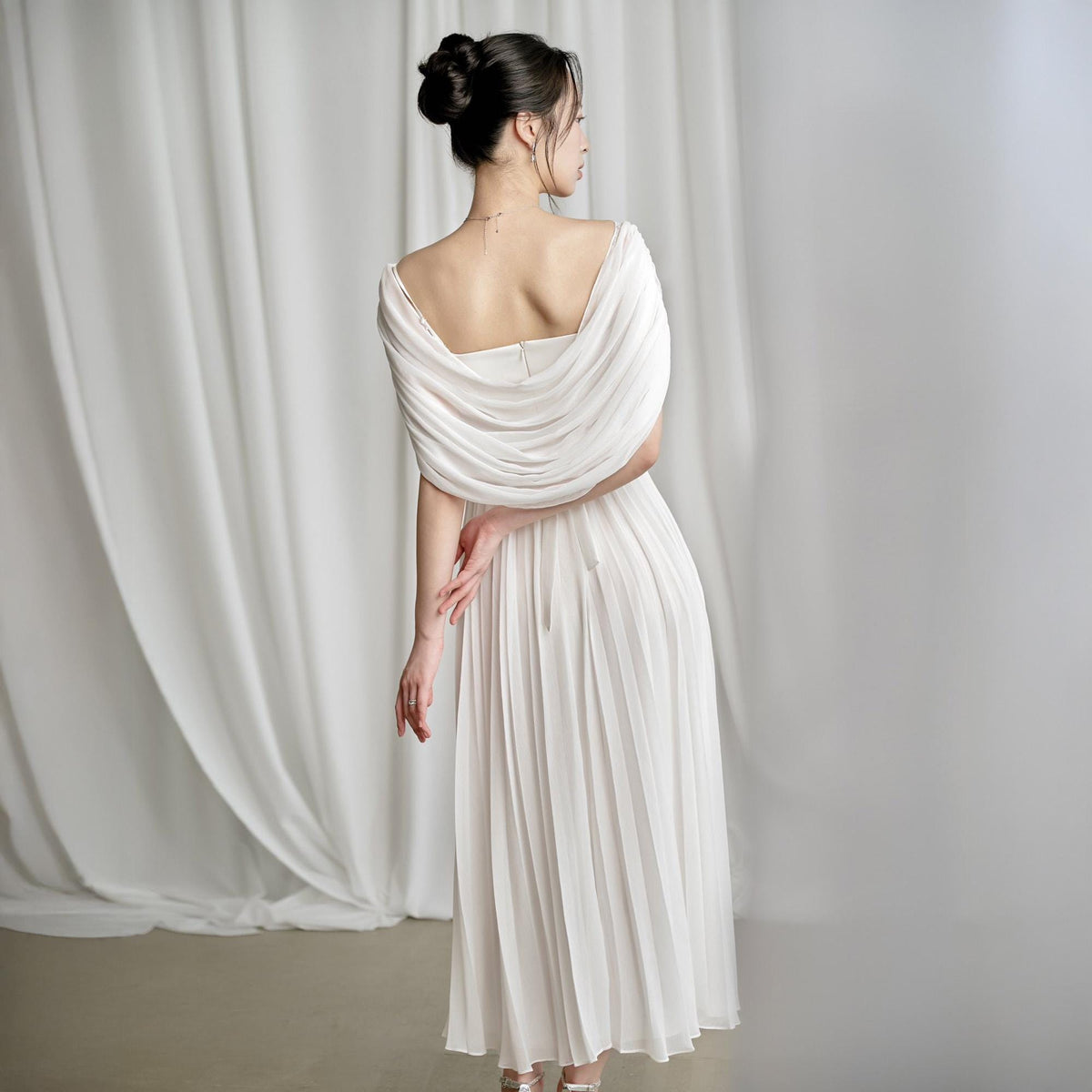 May include: A white, pleated midi dress with a draped, off-the-shoulder neckline. The dress features a flowing, elegant design, perfect for formal events. The model is seen from the back, showcasing the dress's details against a white backdrop.