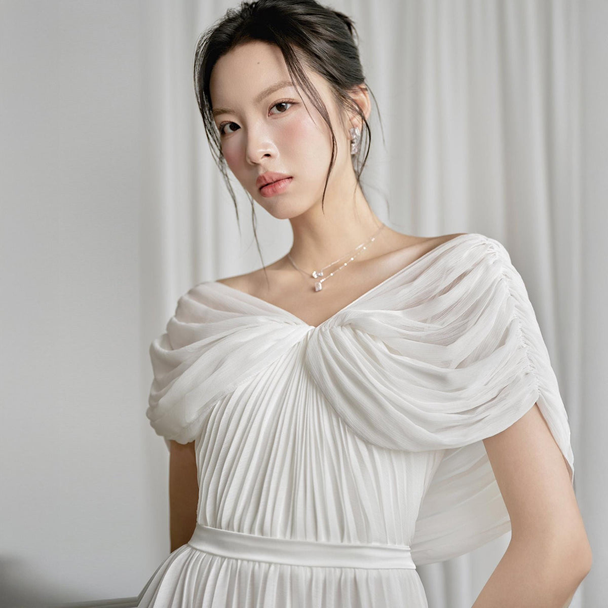 May include: A white, off-the-shoulder dress with a pleated bodice and draped sleeves. The dress features a fitted waist and a delicate necklace. The fabric appears to be lightweight and flowing, creating an elegant silhouette.