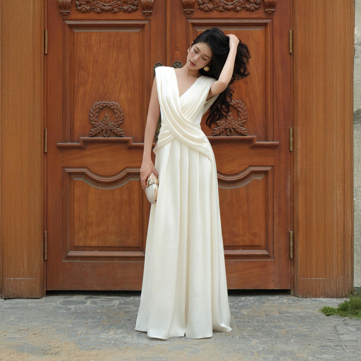 May include: A floor-length ivory gown with a draped bodice and a flowing skirt. The dress features a V-neckline and sleeveless design. A small, gold clutch is held in the hand. The background is a wooden door.