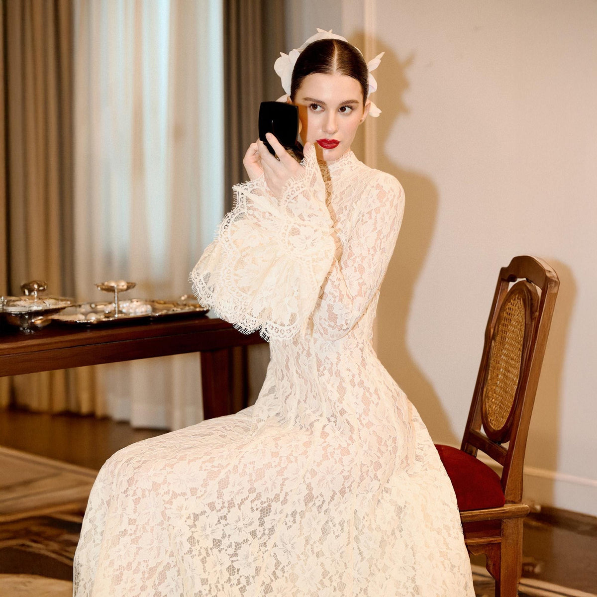 May include: A floor-length cream-colored lace dress featuring a high neckline, long sleeves with flared, scalloped lace cuffs, and a fitted bodice. The garment is crafted from intricate floral lace fabric and is paired with a white floral hair accessory.