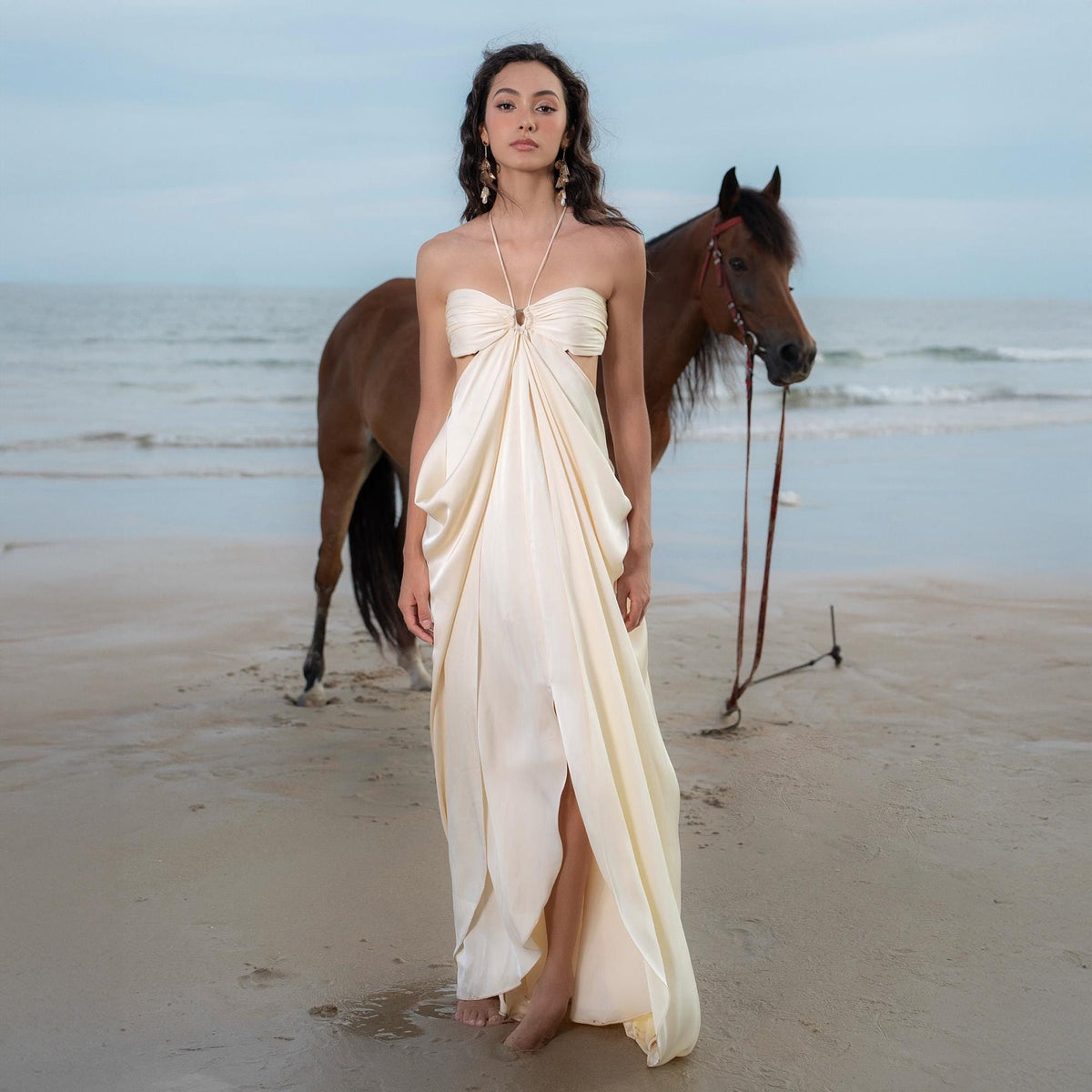 May include: A woman wearing a cream-colored, draped halter dress stands on a beach with a brown horse. The dress features a keyhole cutout and a flowing skirt. The background shows the ocean and a cloudy sky.