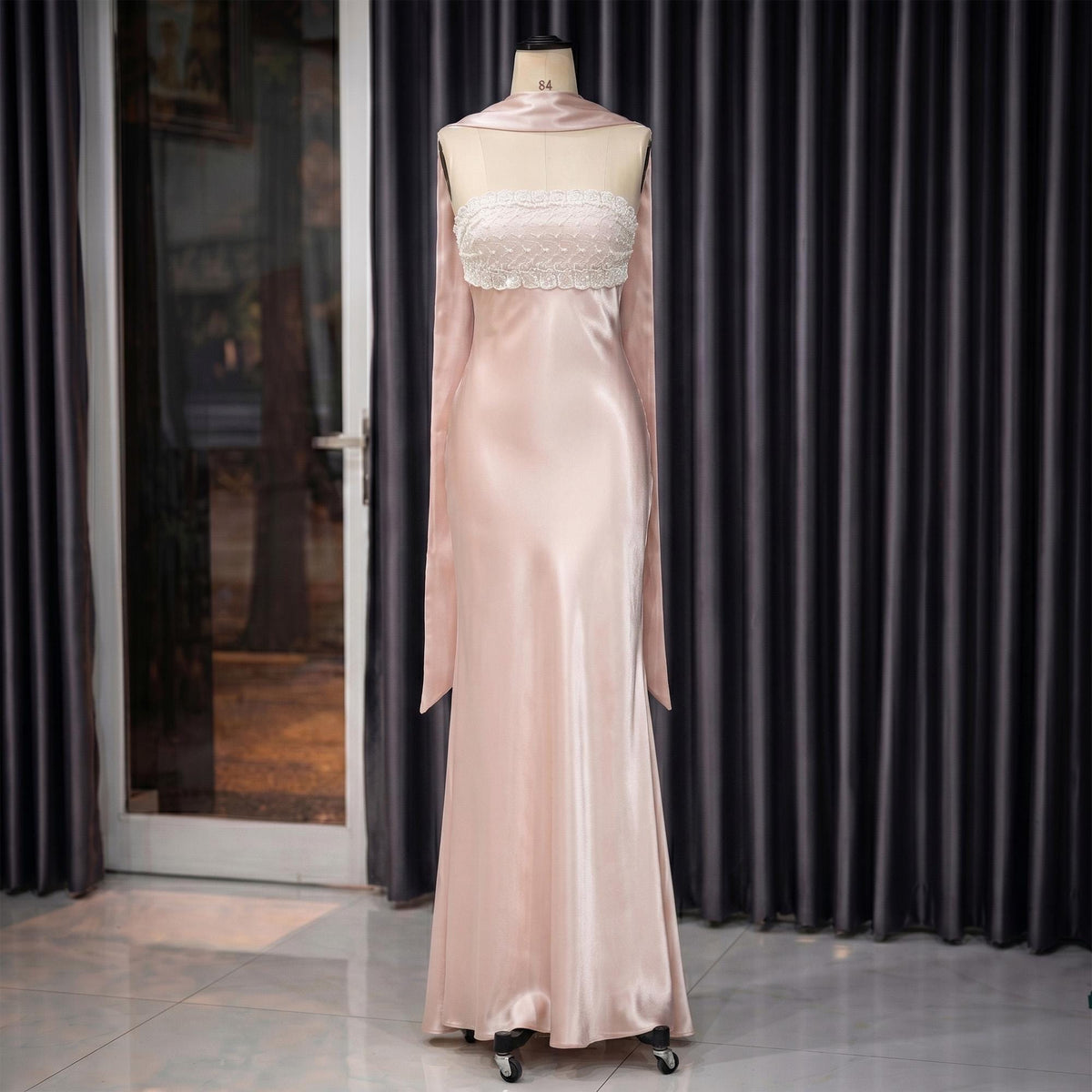 May include: A pale pink satin gown on a mannequin. The dress features a strapless bodice with white lace and a flowing skirt. A matching scarf drapes over the shoulders. The dress is displayed on a rolling stand.