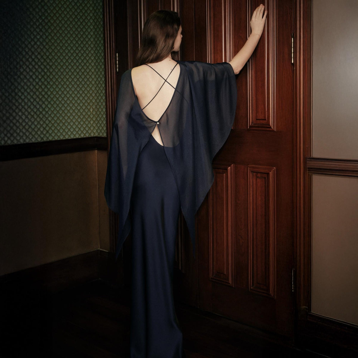 May include: A floor-length dark navy blue dress featuring a sheer, flowing cape-style overlay with wide sleeves. The back of the garment showcases a deep open design with thin, crisscrossing straps that meet at a single central point.