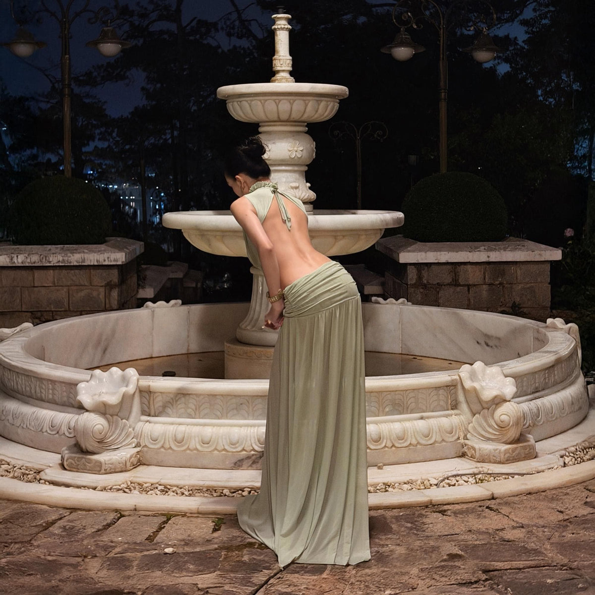 May include: A floor-length sage green dress featuring a halter neckline with thin straps, an open back design, and a gathered waistline that flows into a long, loose skirt. The garment is light in color and has a soft, draped appearance.