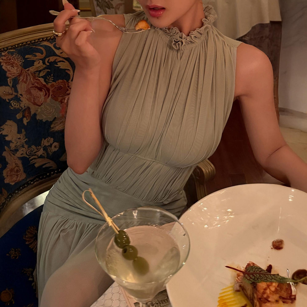 May include: A sleeveless sage green dress featuring a high ruffled neckline and vertical pleated detailing throughout the bodice. The garment has a fitted waistline that transitions into a flowing skirt, creating a structured yet soft silhouette.