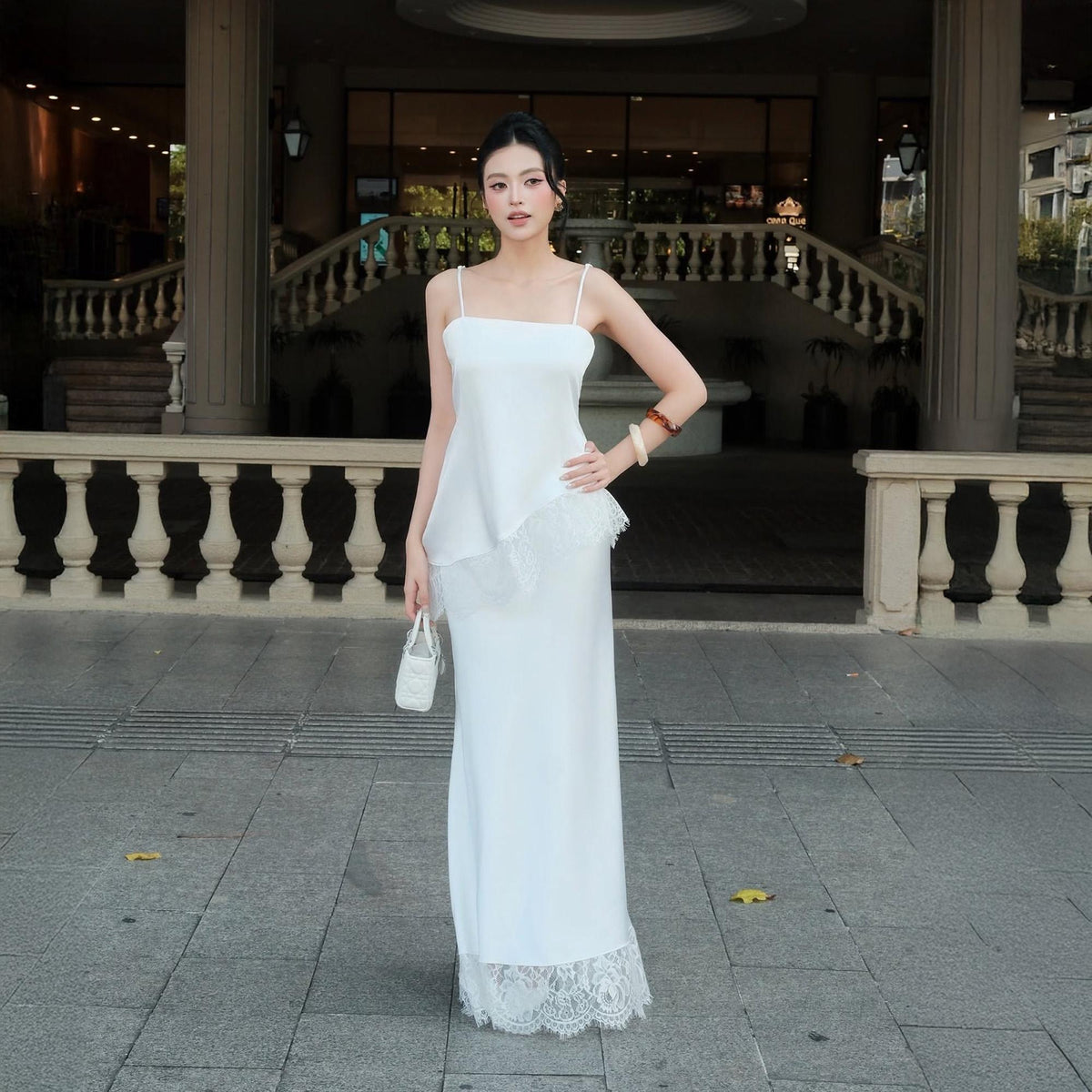 May include: A floor-length white dress with thin spaghetti straps, featuring an asymmetrical layered bodice and a delicate lace trim along the hemline. The garment is paired with a small white handbag and two bracelets on the left wrist.