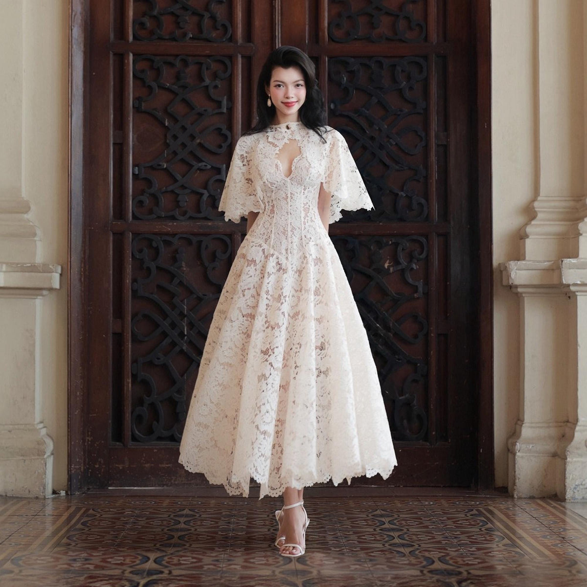 May include: A cream-colored lace midi dress featuring a high neckline with a central cutout, short cape-style sleeves, and a fitted bodice that transitions into a full, flared skirt. The intricate floral lace pattern covers the entire garment.