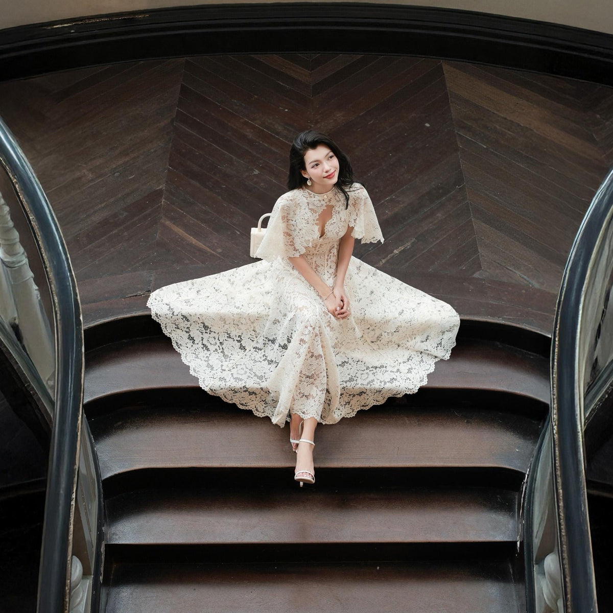 May include: A floor-length white lace dress featuring short flutter sleeves, a high neckline with a small cutout detail, and a scalloped hemline. The dress is crafted from intricate floral lace fabric and is paired with simple white strappy sandals.