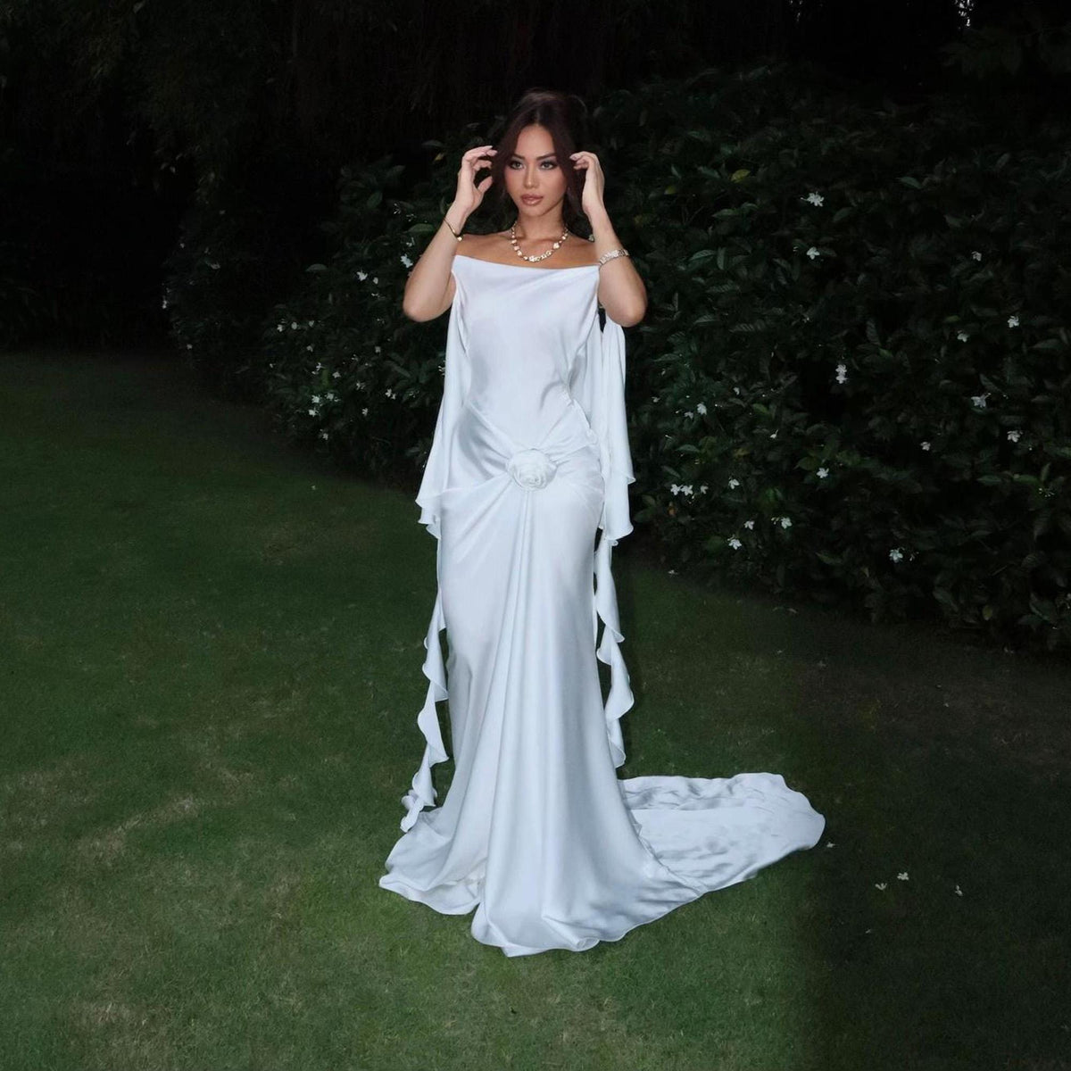 May include: A floor-length white satin gown featuring an off-the-shoulder neckline, draped fabric detailing at the waist with a central circular accent, and flowing side panels that extend into a train, creating a structured yet fluid silhouette.